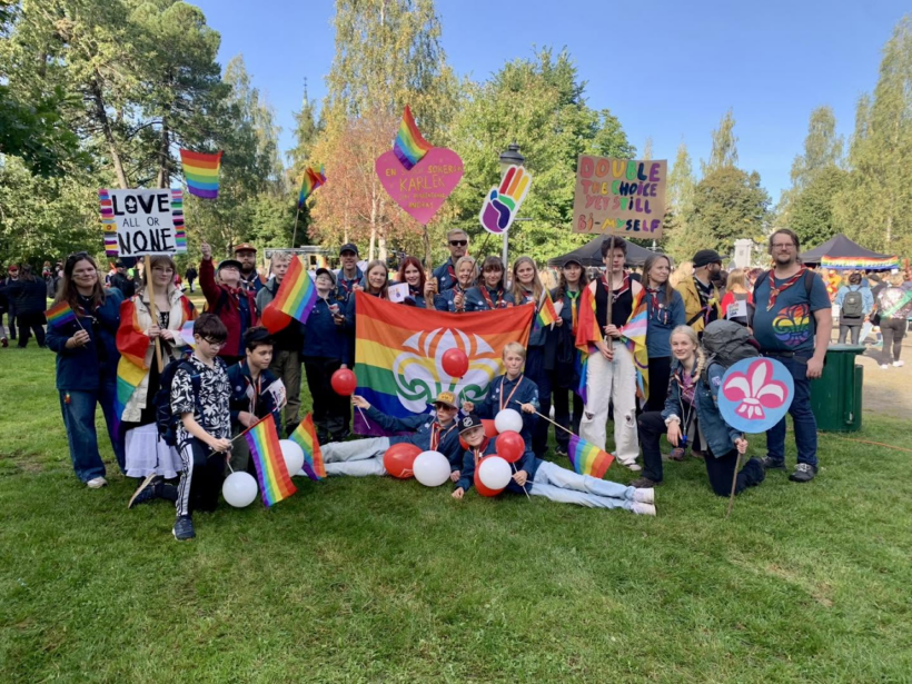 En grupp människor står utomhus på en gräsmatta med regnbågsflaggor, ballonger och skyltar. Skyltarna innehåller budskap som "LOVE ALL OR NONE" och "DOUBLE THE CHOICE YET STILL BI-MYSELF", och flera personer bär scoutuniformer och regnbågsfärgade accessoarer.