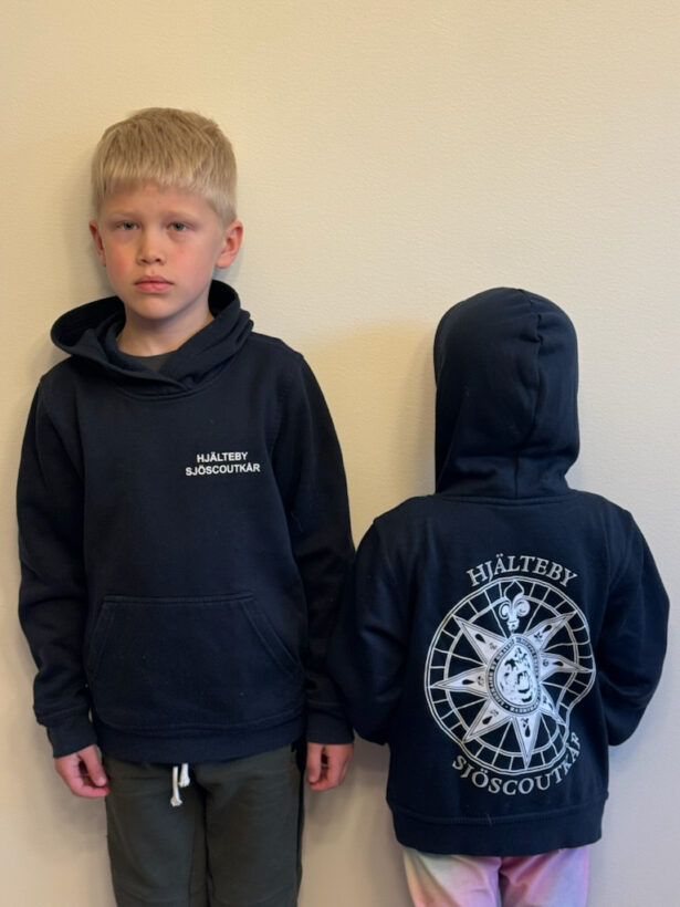 Två barn iförda mörkblå huvtröjor står mot en ljus vägg; den vänstra visar framsidan med texten "Hjälteby Sjöscoutkår" på bröstet, och den högra visar ryggen med ett stort kompassmotiv och samma text.
