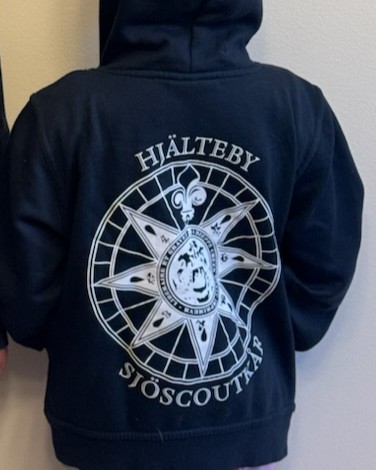 En person i en mörkblå huvtröja med texten "Hjälteby Sjöscoutkår" och en stor kompassros tryckt på ryggen.