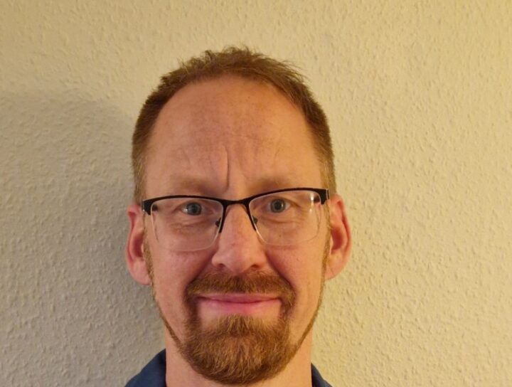 En person klädd i en mörkblå scoutskjorta med märken och en gul-röd-grön halsduk knuten runt halsen. Bakgrunden är en ljus vägg.