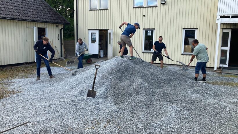 Sex personer arbetar tillsammans med att jämna ut en hög av grus framför en gul byggnad med flera fönster och dörrar. En spade står lutad mot grushögen, och en skottkärra syns i bakgrunden.