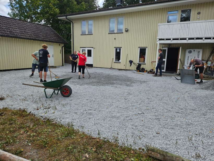 Flera personer arbetar utomhus på en gårdsplan täckt av grus framför en gul träbyggnad; en skottkärra och olika verktyg syns i förgrunden.