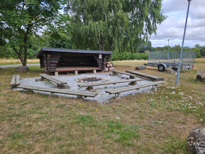 Ett vindskydd av trä står på en grusbelagd yta, omgiven av bänkar och en eldstad i mitten, med grönska, en släpvagn och en lyktstolpe i bakgrunden.