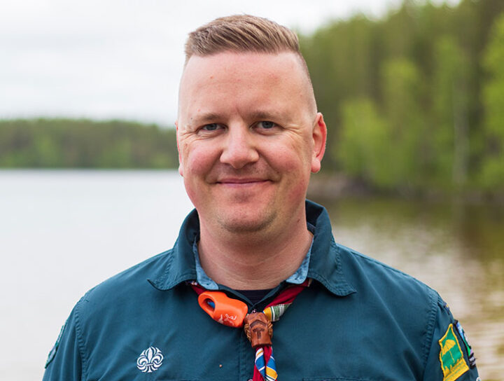 En scout med mörkblå skjorta och halsduk står vid en sjö med skog i bakgrunden.