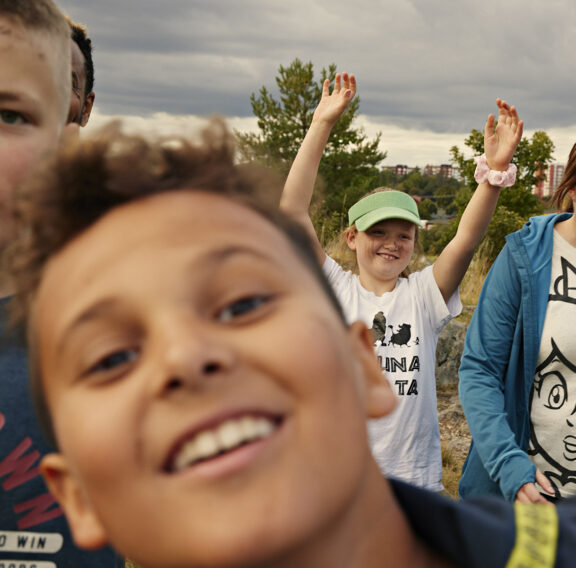 En grupp människor utomhus, med ett barn längst fram som ler mot kameran, ett annat barn i bakgrunden har händerna uppsträckta, och en vuxen kvinna går bredvid med en blå jacka och en t-shirt med en tecknad figur på.