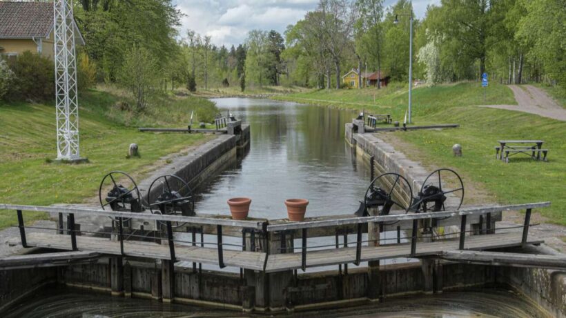 En gammaldags sluss med två svarta vevhjul och träplattformar, omgiven av grönska och en grusväg. I bakgrunden syns ett gult hus och ett rött hus bland träd.