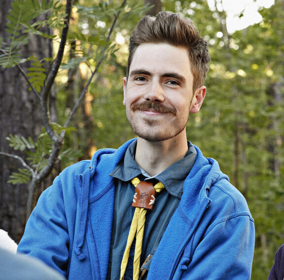 En grupp människor står i skogen och pratar. En person i en blå tröja bär en scoutslips med en knut i trä och gul halsduk.