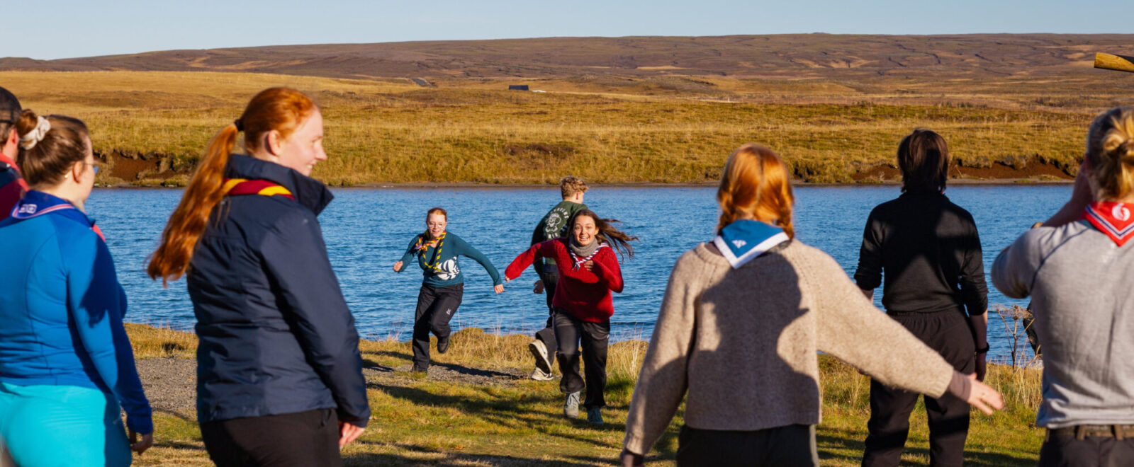 A group of people wearing scout uniforms stands near a lake, watching as two individuals run towards them on a grassy field under a clear blue sky.