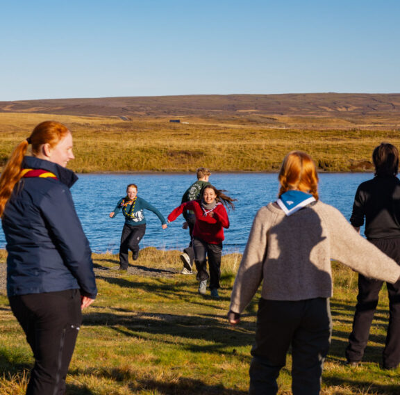 A group of people wearing scout uniforms stands near a lake, watching as two individuals run towards them on a grassy field under a clear blue sky.