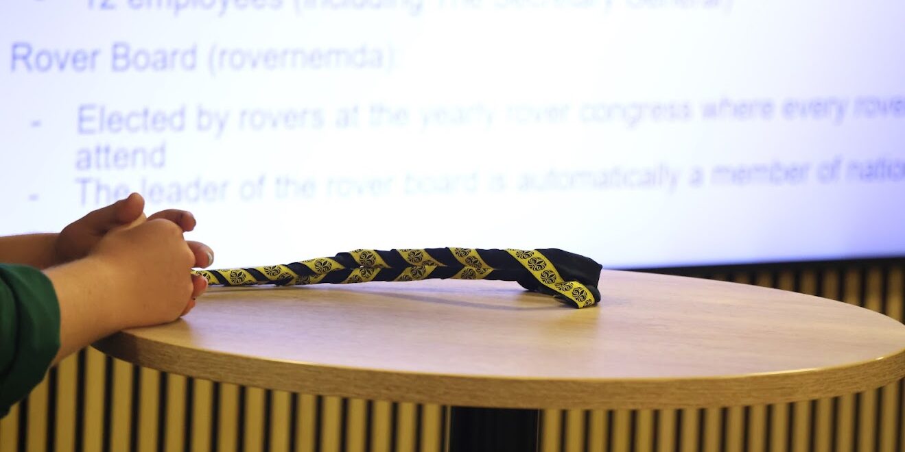 A pair of hands rests on a circular wooden table next to a black and yellow patterned neckerchief, with a blurred presentation slide in the background displaying text about the Secretary General and Rover Board.