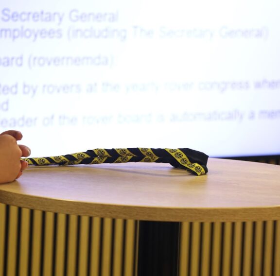 A pair of hands rests on a circular wooden table next to a black and yellow patterned neckerchief, with a blurred presentation slide in the background displaying text about the Secretary General and Rover Board.