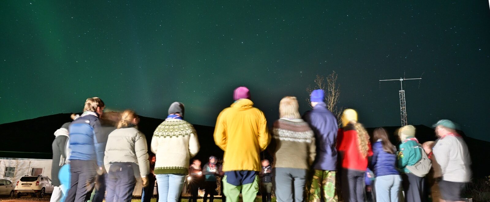 A group of people dressed in warm clothing stand in a circle on a grassy field under a night sky filled with stars and faint green aurora. A radio antenna and a few buildings are visible in the background.