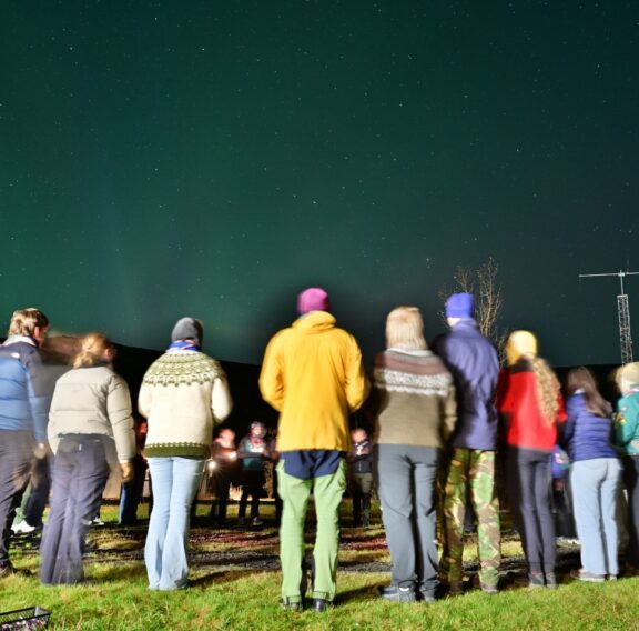 A group of people dressed in warm clothing stand in a circle on a grassy field under a night sky filled with stars and faint green aurora. A radio antenna and a few buildings are visible in the background.