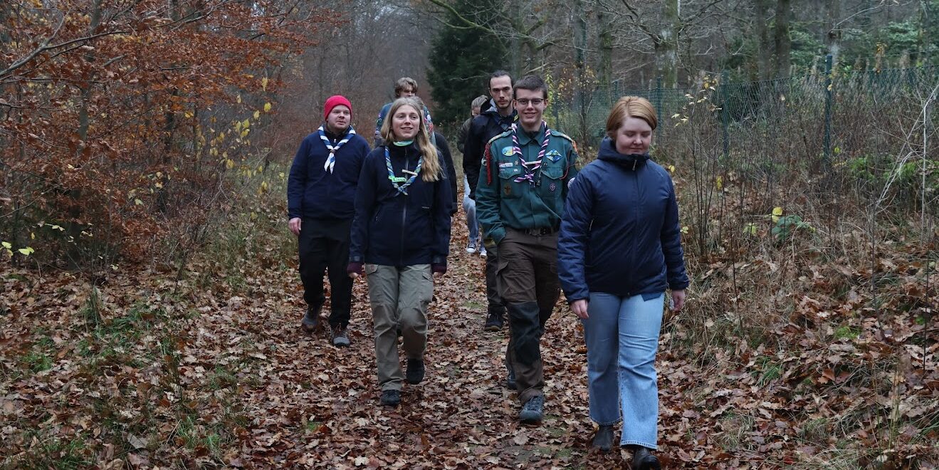 A group of people wearing outdoor clothing and scout neckerchiefs walking along a forest trail covered with autumn leaves, surrounded by bare trees and sparse foliage.