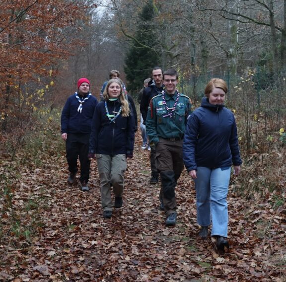 A group of people wearing outdoor clothing and scout neckerchiefs walking along a forest trail covered with autumn leaves, surrounded by bare trees and sparse foliage.