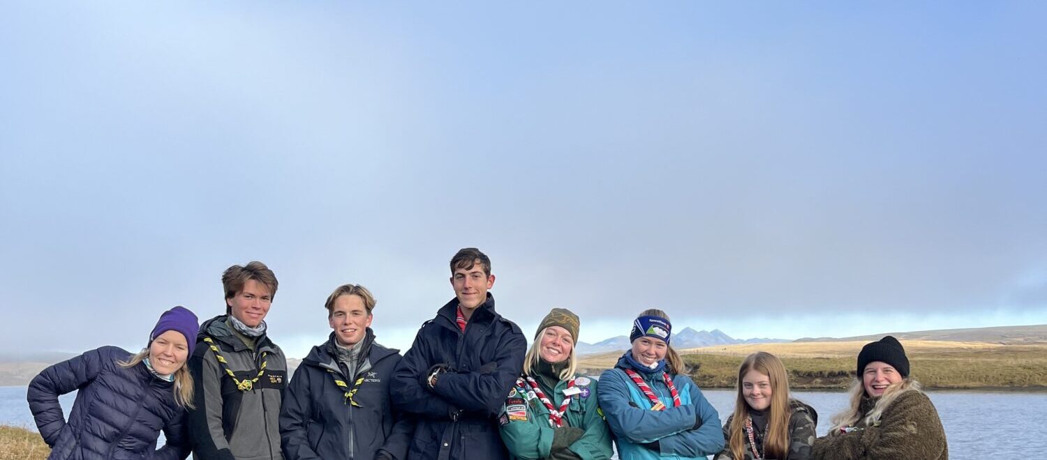 A group of eight people wearing outdoor clothing and scout scarves stand on a grassy area near a body of water, posing casually with a bright blue sky in the background.
