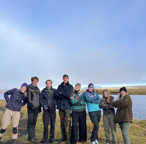 A group of eight people wearing outdoor clothing and scout scarves stand on a grassy area near a body of water, posing casually with a bright blue sky in the background.