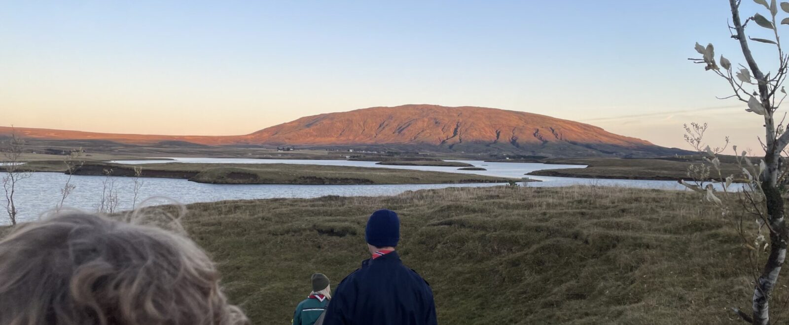 A group of people walks down a grassy hill toward a winding river under a clear blue sky, with a sunlit mountain in the background and sparse trees framing the scene.