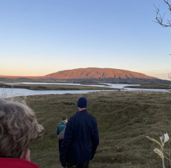 A group of people walks down a grassy hill toward a winding river under a clear blue sky, with a sunlit mountain in the background and sparse trees framing the scene.