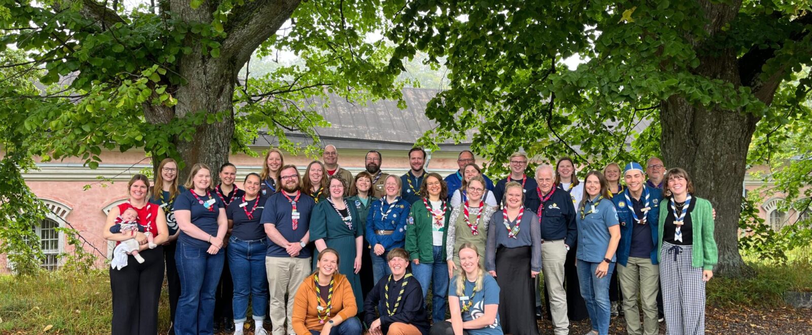 A group of people wearing scout uniforms and neckerchiefs pose under a large tree with green foliage. A brick building with arched windows is visible in the background.