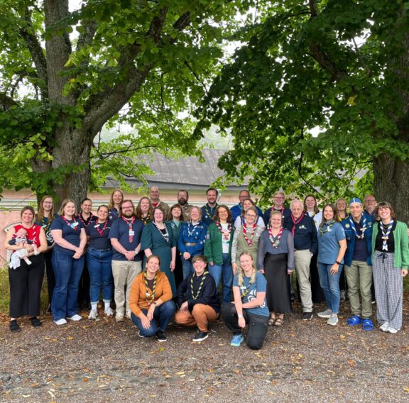 A group of people wearing scout uniforms and neckerchiefs pose under a large tree with green foliage. A brick building with arched windows is visible in the background.