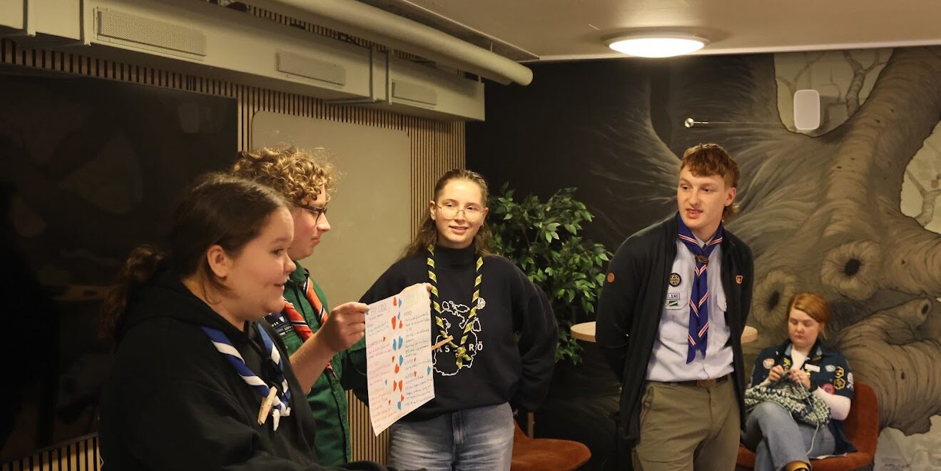 A group of people wearing scout uniforms stand indoors, with one person holding a colorful handwritten paper. The background features a mural of a tree and a small potted plant.