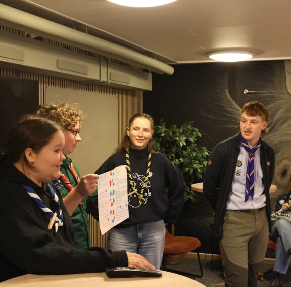 A group of people wearing scout uniforms stand indoors, with one person holding a colorful handwritten paper. The background features a mural of a tree and a small potted plant.