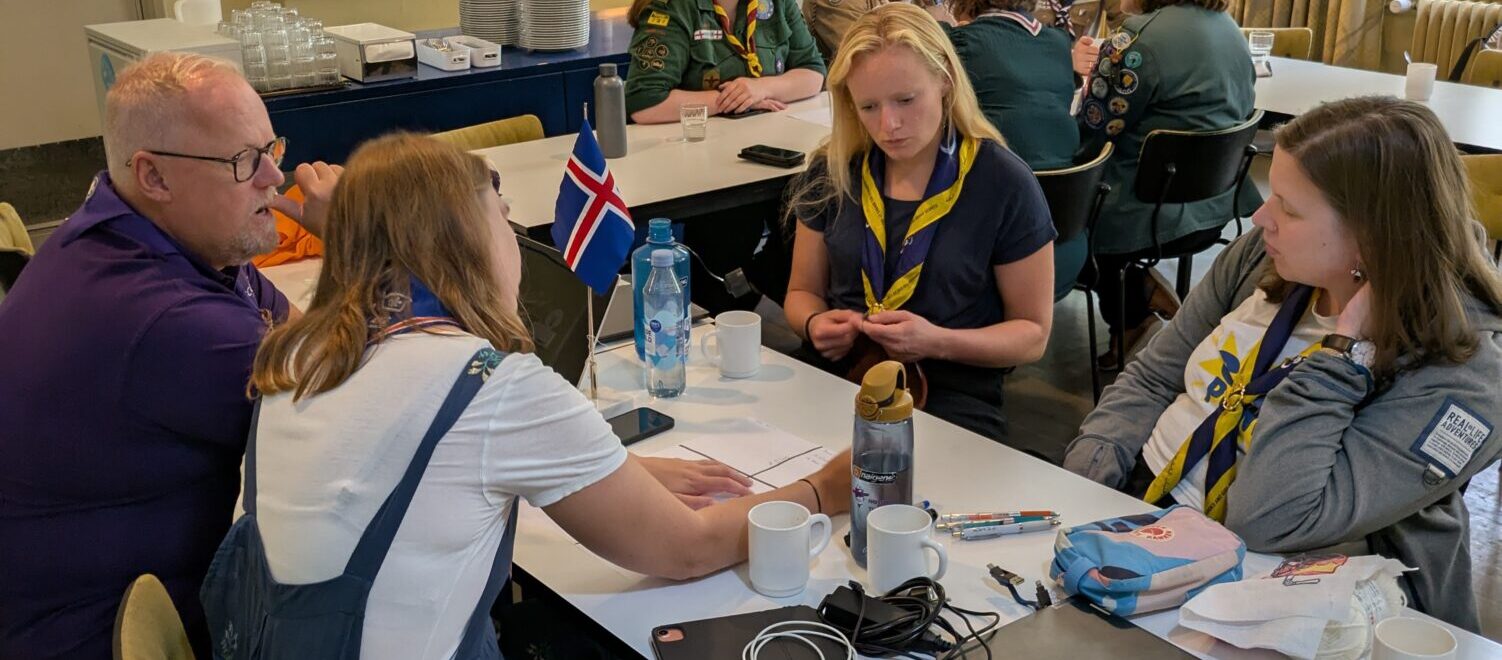 A group of people sit around a table in a casual meeting setting. Some are wearing scout uniforms and scarves, with laptops, notebooks, and cups on the table. An Icelandic flag is visible, and a kitchen area is in the background.