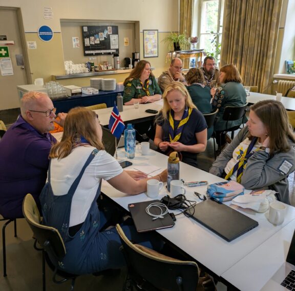 A group of people sit around a table in a casual meeting setting. Some are wearing scout uniforms and scarves, with laptops, notebooks, and cups on the table. An Icelandic flag is visible, and a kitchen area is in the background.