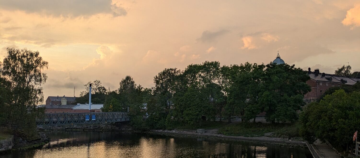 A serene view of a calm waterfront at sunset, with a white wooden railing in the foreground, trees and buildings reflected in the water, and a soft orange and pink sky in the background.
