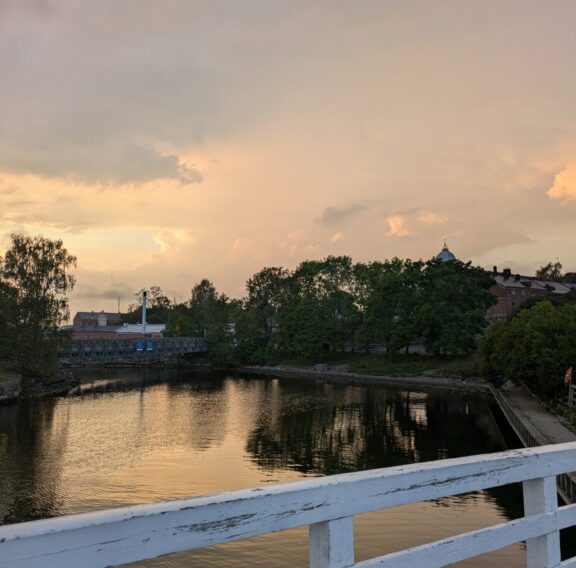 A serene view of a calm waterfront at sunset, with a white wooden railing in the foreground, trees and buildings reflected in the water, and a soft orange and pink sky in the background.