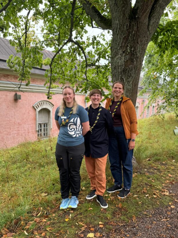 Three people stand together under a tree, with a pink building featuring arched windows in the background. The ground is covered in grass and scattered autumn leaves.